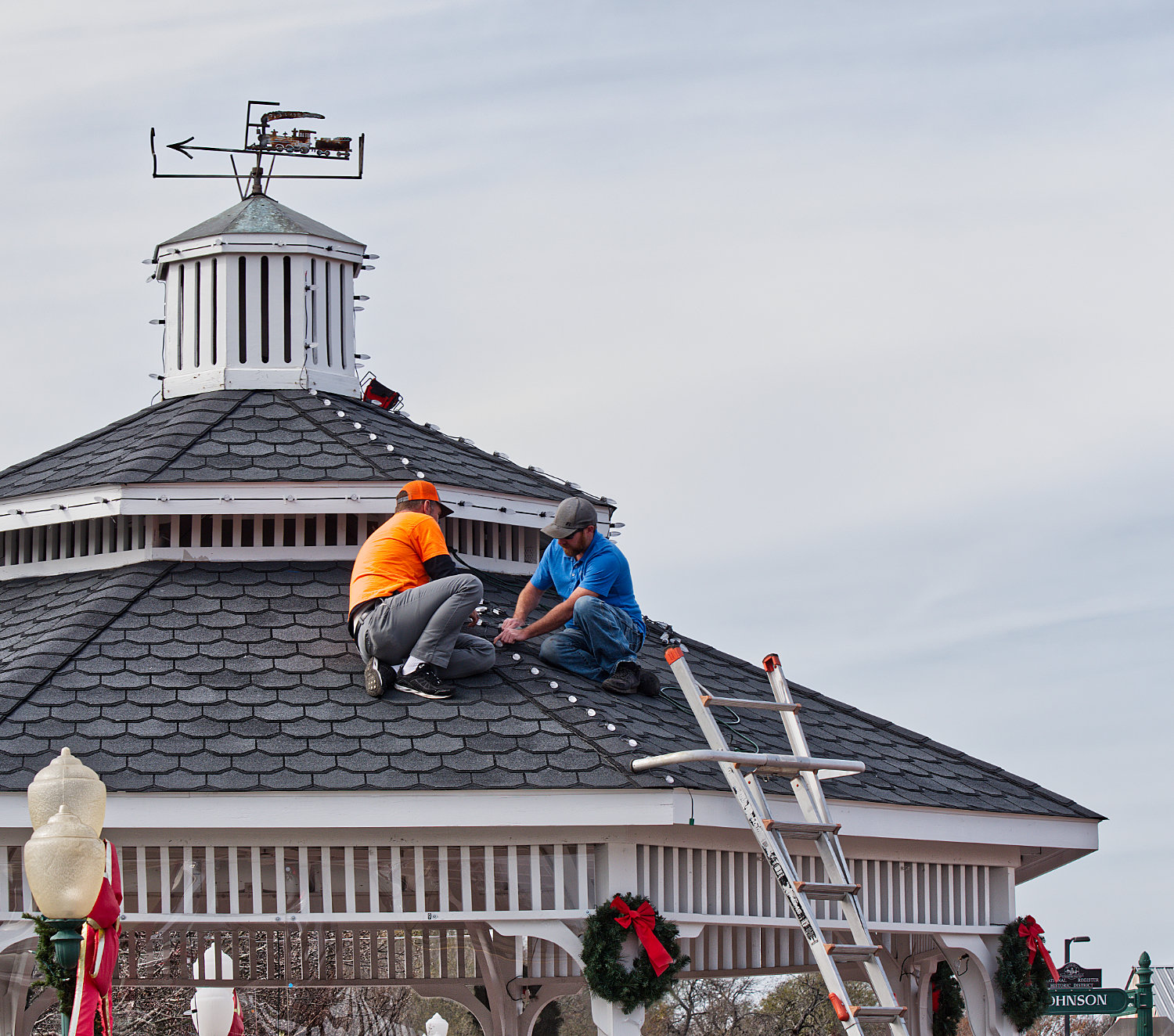 Loyd Hughui (in orange) and Adam Thurman on Thursday string lights along the roof of the gazebo where Santa is expected to appear in downtown Mineola this weekend.